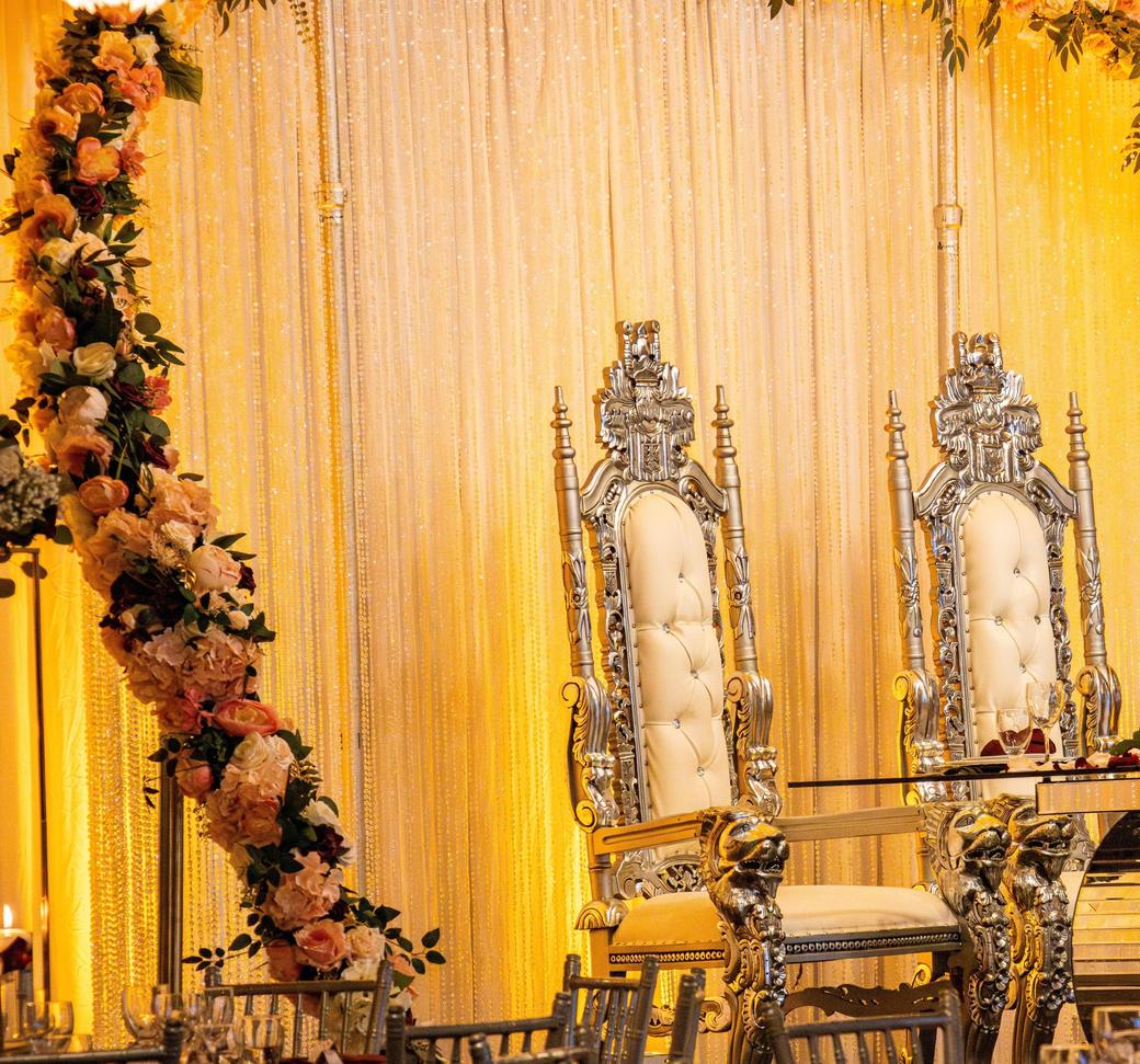 Boda: sillas trono plateadas y arco de flores sobre cortina iluminada